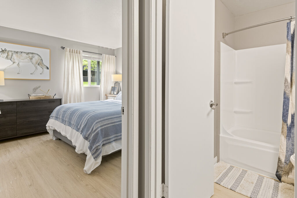 A bright bedroom with a striped blue bedspread and wolf artwork on the wall is shown next to a bathroom with a white bathtub and a patterned shower curtain. Light wood floors run throughout both rooms.