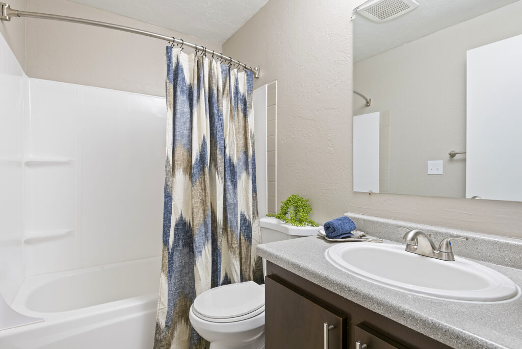 A clean bathroom with a white bathtub and patterned blue and beige shower curtain, a toilet, gray countertop with sink, silver faucet, towel, potted plant, and a large wall mirror above the sink.