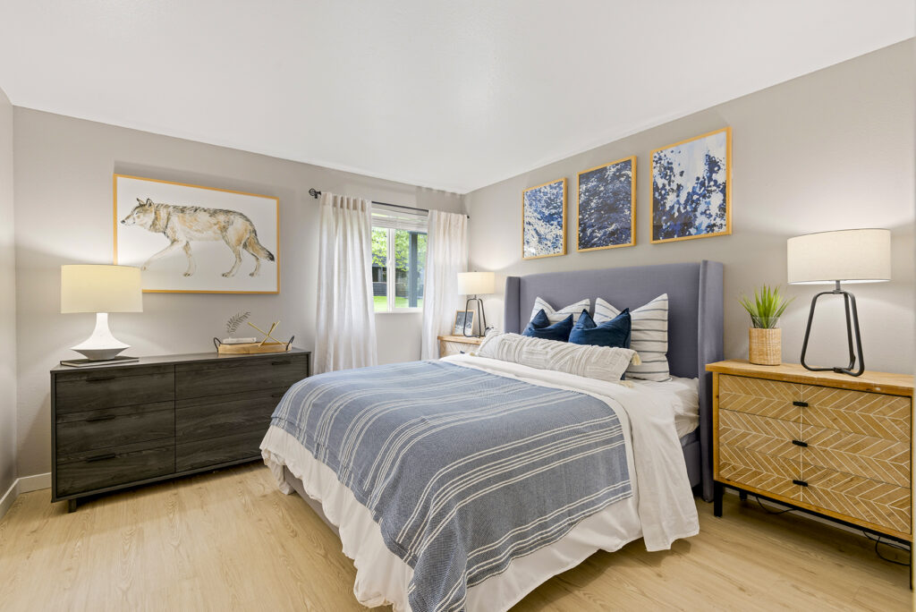 A cozy bedroom with a blue and white striped bed, two nightstands with lamps, a dresser, modern decor, and art on the walls, including a wolf print and three blue abstract pieces above the bed. Light wood flooring and a window with curtains.