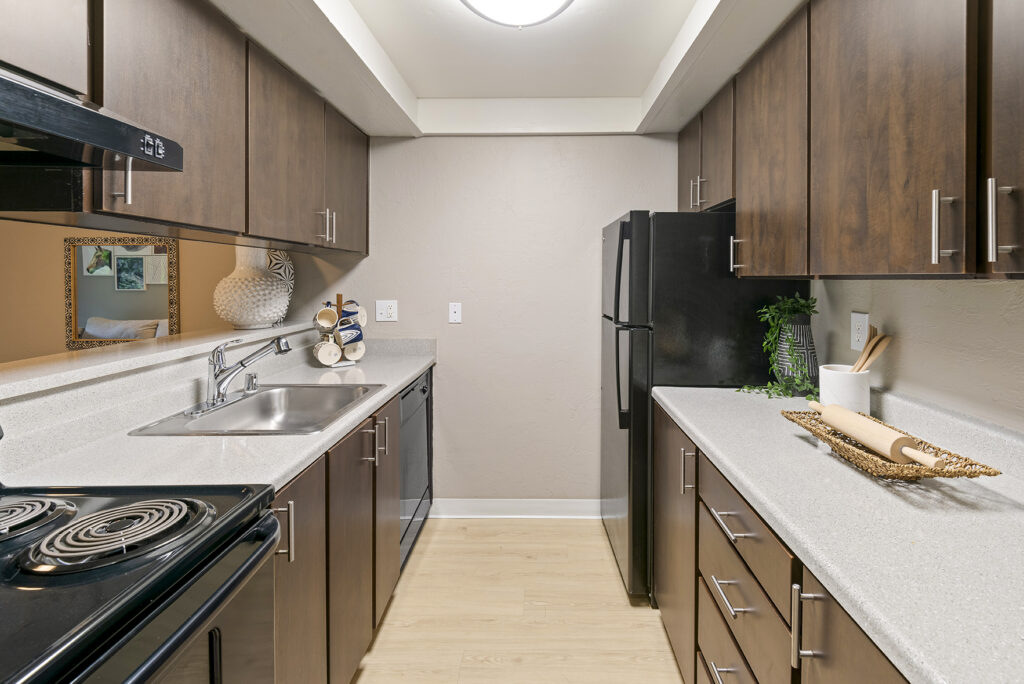 Modern galley kitchen with dark wood cabinets, light countertops, black appliances, and light wood flooring. Sink and stove are on the left; refrigerator and decorative items are on the right. Bright lighting above.