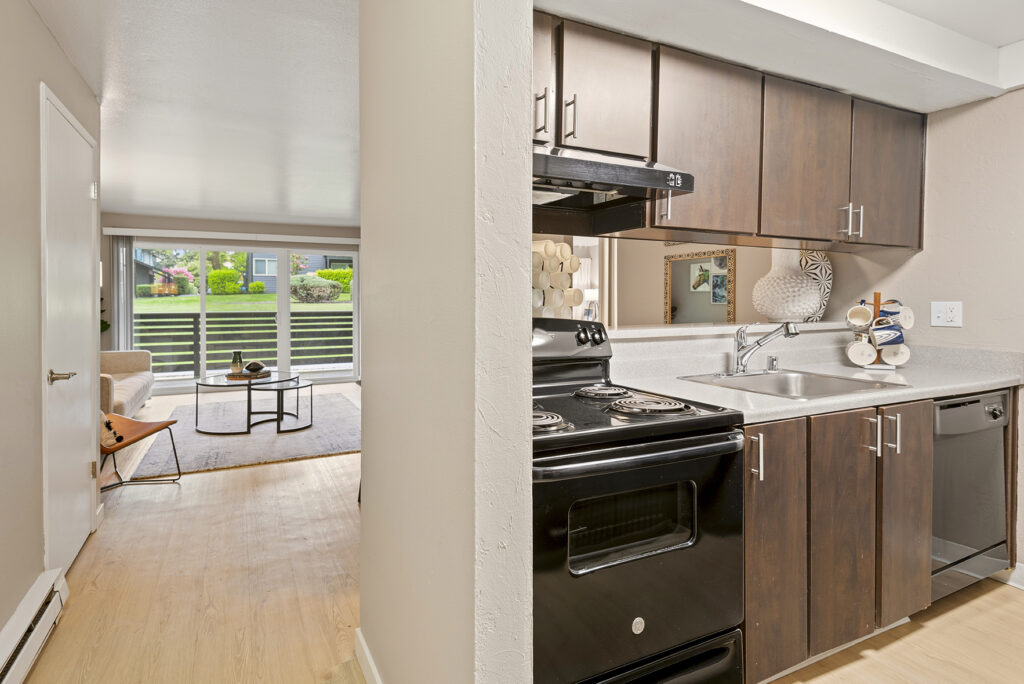 A modern apartment kitchen with dark wood cabinets and black appliances opens to a bright living room with large windows, light wood flooring, and a beige sofa. A glass coffee table is centered on a neutral rug.