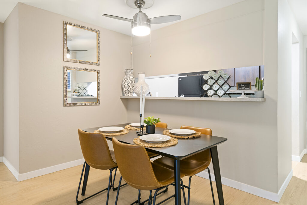 A modern dining area with a black table, four tan leather chairs, place settings, and a small plant centerpiece. Two decorative mirrors hang on the wall, and a kitchen is visible through a pass-through window.