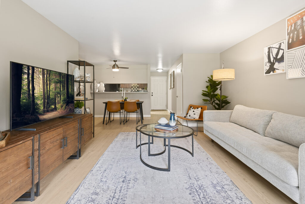 Modern living room with a light gray sofa, round glass coffee table, wooden TV stand with a large TV, dining area with four chairs, open kitchen, and wall art. Neutral tones and a potted plant create a cozy atmosphere.